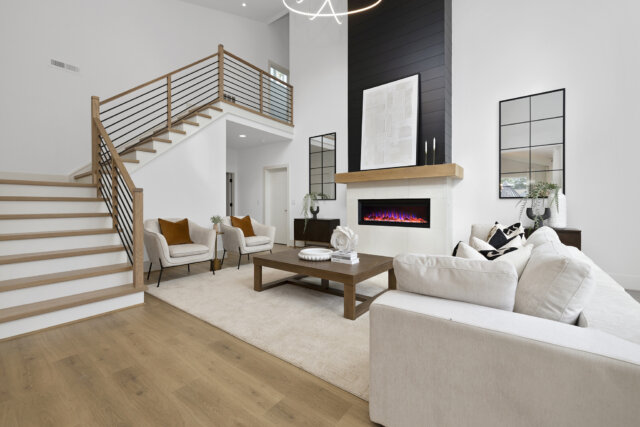 Modern living room with a double-height ceiling, featuring a sleek electric fireplace with a wood mantel and black shiplap accent wall. The space includes a neutral-toned sectional sofa, mid-century armchairs, a wood coffee table, and a light wood staircase with horizontal metal railing. Natural light fills the room through large windows, highlighting the clean lines and minimalist decor.